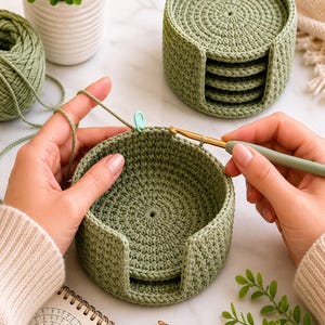 May include: A sage green crocheted basket with a matching set of circular coasters inside. The basket features a thick, textured stitch pattern with a U-shaped cutout on the front. A metal crochet hook is actively working on the basket rim.