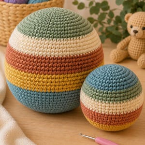 May include: Two spherical crochet balls featuring horizontal stripes in sage green, cream, terracotta, yellow, and teal. The larger ball is positioned behind the smaller one, both crafted with a consistent tight stitch pattern in a multi-colored design.