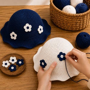 May include: A dark blue crochet bucket hat with white flower appliqués and a white crochet bucket hat with dark blue flower appliqués. A needle is used to attach a dark blue flower to the white hat, with extra yarn and flower pieces nearby.
