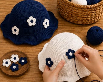 May include: A dark blue crochet bucket hat with white flower appliqués and a white crochet bucket hat with dark blue flower appliqués. A needle is used to attach a dark blue flower to the white hat, with extra yarn and flower pieces nearby.