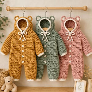 May include: Three long-sleeved crochet rompers with hoods featuring bear ears. The rompers are mustard yellow, sage green, and dusty pink. Each has a white trim, a white drawstring tie at the neck, and a vertical row of white buttons down the front.