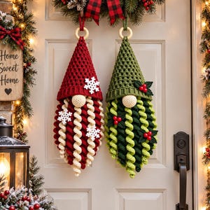 May include: Two hanging crochet gnome decorations with pointed hats and long curly beards. One gnome features a red hat with white snowflake accents, while the other has a green hat with holly leaf and red berry embellishments on a wooden ring.