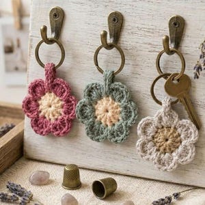 May include: Three crocheted flower keychains in pink, sage green, and cream, each featuring a central circle and six petals. The keychains are attached to bronze-colored metal rings and hung on matching bronze hooks. One key is attached to the cream keychain.
