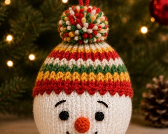 May include: A round, white crocheted snowman ornament featuring a knitted hat with red, yellow, and green horizontal stripes, topped with a multicolored pom-pom and a red hanging loop. The face has black embroidered eyes, eyebrows, and a smile.