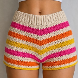May include: Crocheted shorts with a vibrant, striped pattern. The shorts feature alternating bands of cream, pink, orange, and yellow. The waistband is a solid cream color. The shorts are a high-waisted style.