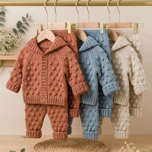 May include: Three sets of knitted baby clothing hanging on wooden hangers. Each set includes a hooded cardigan with buttons and matching trousers, featuring a textured bobble pattern. The sets are available in terracotta, dusty blue, and cream colours.