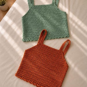 Può includere: Due top all'uncinetto fatti a mano. Uno è verde salvia, l'altro arancione bruciato. Entrambi hanno scollature quadrate, bordi smerlati e spalline larghe. I top sono esposti su una superficie bianca.