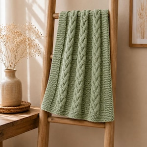 May include: A sage green knitted blanket featuring a prominent vertical cable knit pattern. The blanket has a textured border and is draped over a wooden ladder. The knit construction displays uniform stitches and a soft, matte finish throughout.