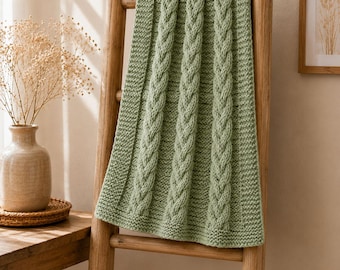 May include: A sage green knitted blanket featuring a prominent vertical cable knit pattern. The blanket has a textured border and is draped over a wooden ladder. The knit construction displays uniform stitches and a soft, matte finish throughout.