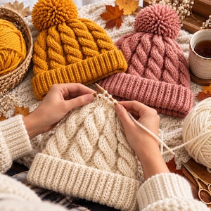 May include: Three knitted beanies featuring a cable knit pattern and a large pom-pom on top. One beanie is mustard yellow, one is dusty rose, and one is cream-coloured. The cream beanie is currently being worked on with wooden knitting needles.