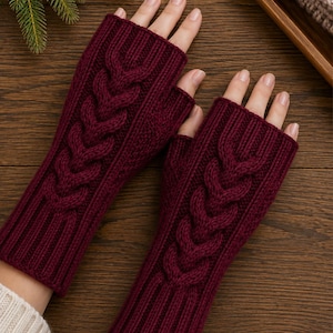 May include: A pair of deep burgundy fingerless gloves featuring a prominent vertical cable knit pattern running down the center. The gloves have a ribbed cuff at the wrist and a ribbed opening for the fingers, crafted from a thick, textured knit fabric.