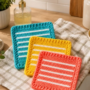 May include: Three colorful, square crochet washcloths in turquoise, yellow, and coral, each with white horizontal stripes. The washcloths are displayed on a checkered cloth, suggesting a kitchen or bathroom setting.