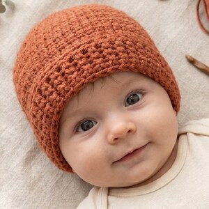 May include: A crocheted rust-colored baby beanie. The hat is made of a textured yarn and has a folded brim. The baby is wearing a cream-colored onesie. A wooden crochet hook and a ball of yarn are in the background.