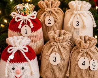 May include: A collection of six knitted drawstring pouches for an advent calendar. One pouch features a Santa face design, while others are solid beige or white. Each pouch is tied with a white cord and includes a round wooden tag numbered 1 through 4.