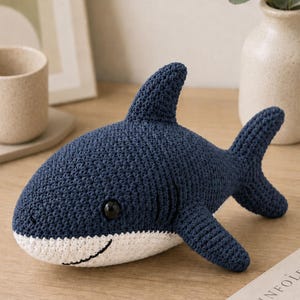 Peut inclure: Un jouet en peluche requin crocheté bleu foncé et blanc avec une nageoire dorsale proéminente, une nageoire latérale, une queue et une bouche cousue noire. Le requin a un corps arrondi avec un ventre blanc et un œil en plastique noir.