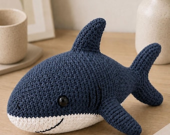 May include: A dark blue and white crocheted shark plush toy featuring a prominent dorsal fin, a side fin, a tail, and a black stitched mouth. The shark has a rounded body with a white underbelly and a single visible black plastic eye.