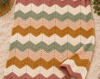 May include: A crocheted blanket featuring a repeating chevron pattern in horizontal stripes of dusty rose, cream, sage green, and mustard yellow. The textile has a textured stitch construction with a scalloped edge along the top and bottom.