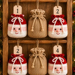 May include: Six knitted drawstring pouches hanging in a wooden frame. Four pouches feature a red, pink, and white face design with a numbered tag. Two pouches are solid tan with a drawstring closure. Each pouch has a jute twine loop for hanging.