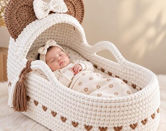 May include: A cream-colored crocheted baby bassinet featuring two large brown circular ears and a central cream bow on the hood. The bassinet is decorated with small brown heart-shaped accents along the base and includes a matching cream tassel.