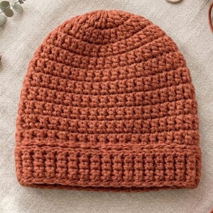 May include: A handmade, rust-colored crochet beanie hat. The hat is made with a thick yarn and features a ribbed brim and a close-knit stitch pattern. The hat is a warm, cozy accessory.