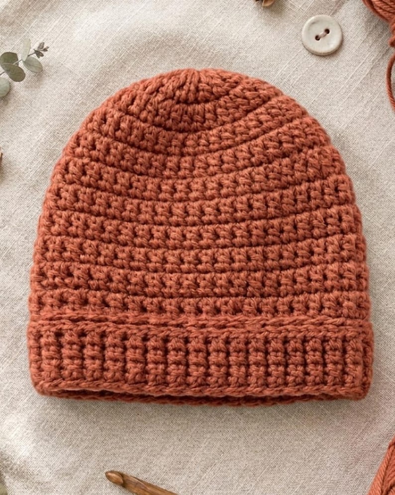 May include: A handmade, rust-colored crochet beanie hat. The hat is made with a thick yarn and features a ribbed brim and a close-knit stitch pattern. The hat is a warm, cozy accessory.