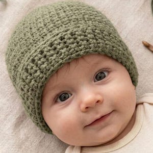 May include: A close-up of a baby wearing a sage green crochet beanie. The baby is wearing a cream-colored onesie. The beanie is handmade with a textured stitch. The baby has dark eyes and a slight smile.
