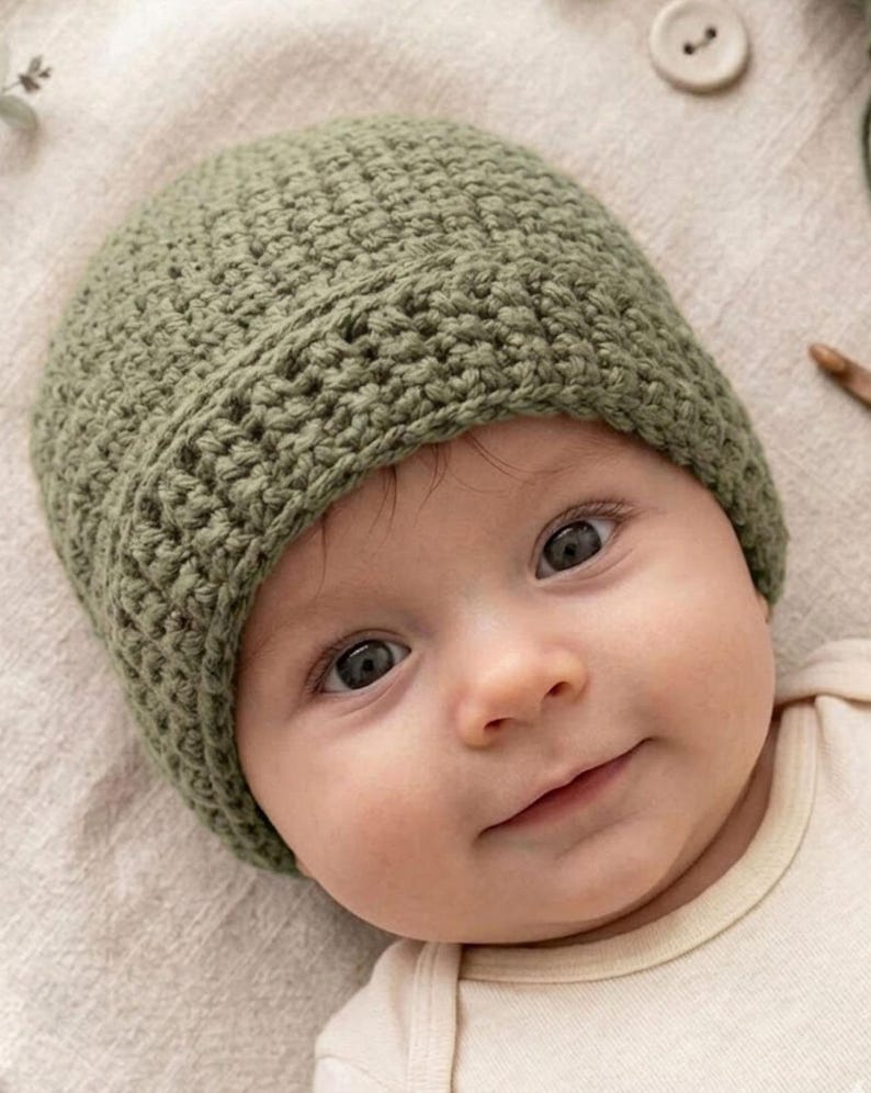May include: A close-up of a baby wearing a sage green crochet beanie. The baby is wearing a cream-colored onesie. The beanie is handmade with a textured stitch. The baby has dark eyes and a slight smile.