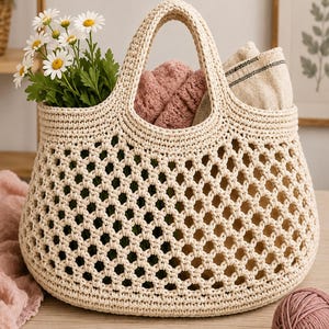 Op de afbeelding: Een crèmekleurige gehaakte tas met een open ruitpatroon, twee stevige handvatten en een dichte bodem. De tas bevat een boeket witte madeliefjes met gele harten, een opgevouwen roze textiel en een beige stof met een zwarte bies.
