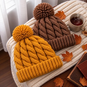 Puede incluir: Dos gorros de punto con pompones, uno de color amarillo mostaza y otro de color marrón oscuro. Ambos gorros presentan un patrón de punto de ochos grueso y un borde acanalado ancho. Están hechos de lana y tienen una textura gruesa.