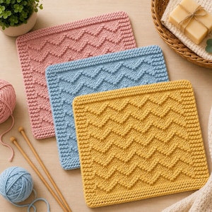 May include: Three square knitted washcloths in pink, light blue, and mustard yellow, each featuring a textured chevron pattern and a garter stitch border. The set includes two balls of yarn, two wooden knitting needles, and a bar of soap.