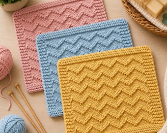 May include: Three square knitted washcloths in pink, light blue, and mustard yellow, each featuring a textured chevron pattern and a garter stitch border. The set includes two balls of yarn, two wooden knitting needles, and a bar of soap.