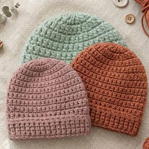 May include: Three handmade crochet beanies in soft pastel colors: dusty rose, sage green, and burnt orange. The hats are arranged on a neutral linen surface, showcasing the textured stitch work. Additional yarn, buttons, and a crochet hook are visible.