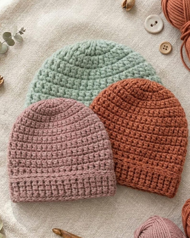 May include: Three handmade crochet beanies in soft pastel colors: dusty rose, sage green, and burnt orange. The hats are arranged on a neutral linen surface, showcasing the textured stitch work. Additional yarn, buttons, and a crochet hook are visible.
