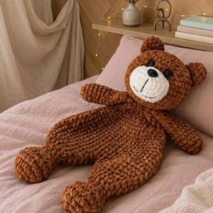 May include: A handmade brown crochet teddy bear with a white face, black eyes, and a stitched smile. The bear is lying on a pink bedspread. The bear's arms and legs are extended.