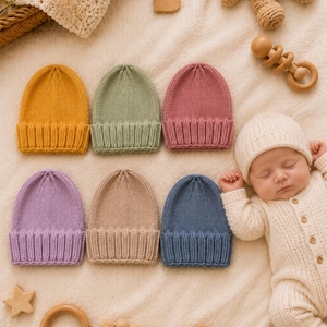 May include: A collection of six knitted baby hats in mustard yellow, sage green, dusty rose, lavender, beige, and slate blue, each featuring a ribbed cuff and a smooth stockinette stitch crown, displayed alongside a sleeping infant in a cream onesie.