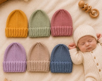 May include: A collection of six knitted baby hats in mustard yellow, sage green, dusty rose, lavender, beige, and slate blue, each featuring a ribbed cuff and a smooth stockinette stitch crown, displayed alongside a sleeping infant in a cream onesie.