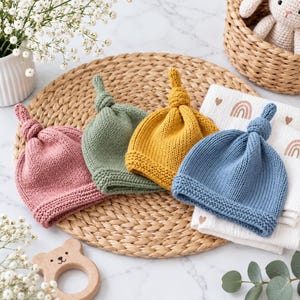 Puede incluir: Cuatro gorros de punto para bebé en rosa empolvado, verde salvia, amarillo mostaza y azul pizarra. Cada gorro presenta una parte superior anudada y un borde texturizado en punto bobo. Los gorros están dispuestos en fila con un diseño sencillo.