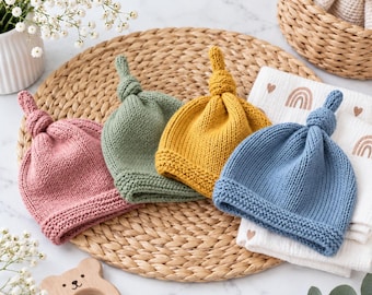 May include: Four knit baby hats in dusty pink, sage green, mustard yellow, and slate blue. Each hat features a knotted top and a textured garter stitch brim. The hats are arranged in a row, showcasing a simple, classic design suitable for infants.