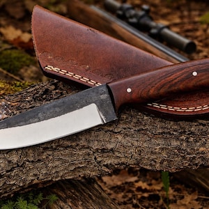 Peut inclure: Un couteau de chasse artisanal avec un manche en bois foncé et un étui en cuir. La lame est argentée et le manche est fixé avec des rivets. L'étui est marron et cousu avec un fil de couleur claire. Le couteau repose sur un morceau de bois.