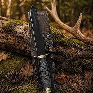 Puede incluir: Un cuchillo de caza negro con mango de madera y detalles dorados está enfundado en una funda de cuero marrón. La funda presenta un diseño de cabeza de ciervo y una correa con un botón dorado. El cuchillo y la funda descansan sobre un tronco cubierto de musgo.