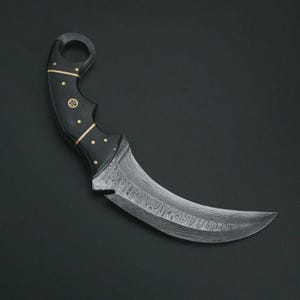 May include: A curved karambit knife with a black handle adorned with gold-colored accents. The blade features a distinctive patterned design. The knife is set against a dark background.