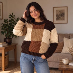 May include: A long-sleeve knit sweater featuring a chunky ribbed texture and a color-blocked pattern in shades of dark brown, medium brown, and cream. The garment has a crew neckline and ribbed cuffs, paired with blue denim jeans.