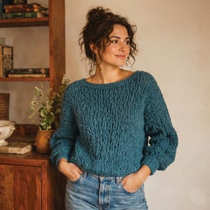 May include: Teal-colored knit sweater with a textured pattern and long sleeves. The sweater has a boat neck and is paired with blue jeans. The image is taken indoors, with a wooden bookshelf and plants in the background.