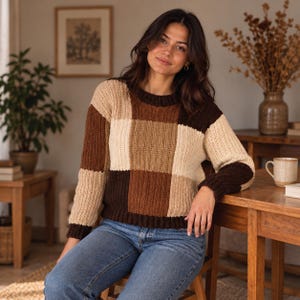 May include: A long-sleeve knit sweater featuring a large color-blocked pattern in shades of dark brown, medium brown, and cream. The garment has a ribbed crew neckline, ribbed cuffs, and a ribbed hem, constructed with a vertical textured stitch.