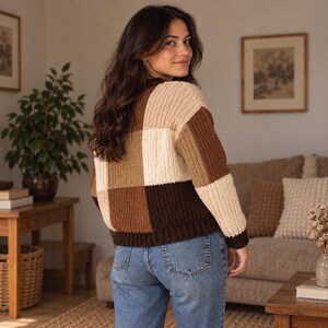 May include: A long-sleeve knit sweater featuring a large checkered pattern in shades of cream, tan, and dark brown. The sweater has a ribbed texture, a crew neckline, and ribbed cuffs and hem, paired with classic blue denim jeans.