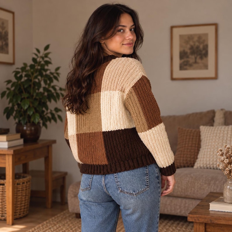 May include: A long-sleeve knit sweater featuring a large checkered pattern in shades of cream, tan, and dark brown. The sweater has a ribbed texture, a crew neckline, and ribbed cuffs and hem, paired with classic blue denim jeans.