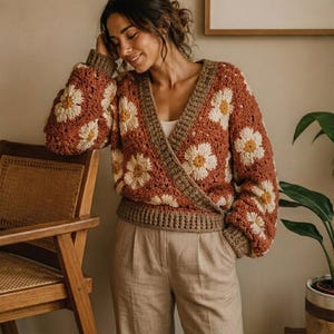 Può includere: Un maglione all'uncinetto color ruggine, stile avvolgente, con motivi a margherita bianchi e gialli. Il maglione ha maniche lunghe e un bordo marrone. La modella indossa pantaloni beige.
