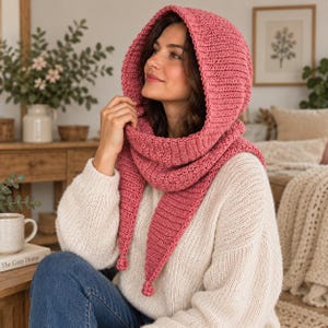 May include: A dusty rose pink crocheted hooded scarf featuring a ribbed texture, a deep hood, and two long pointed ends that hang down the front, each finished with a small decorative knot at the tip, worn over a cream-colored knit sweater.