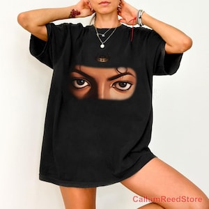 May include: A black short-sleeved t-shirt featuring a graphic print of a pair of eyes and a small crown icon centered on the chest. The design is positioned below the neckline, showcasing a realistic illustration of human eyes in a dark tone.