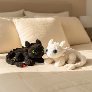 May include: Two crocheted dragon plush toys. One is black with green eyes and a red tip on its tail. The other is white with blue eyes. Both are lying on a bed with cream-colored bedding and pillows.